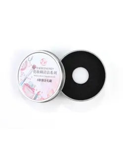Open view of the black circular dry cleaning sponge revealed inside its metal case, with pink-handled makeup brush nearby, demonstrating the sponge's textured surface for quick powder absorption.