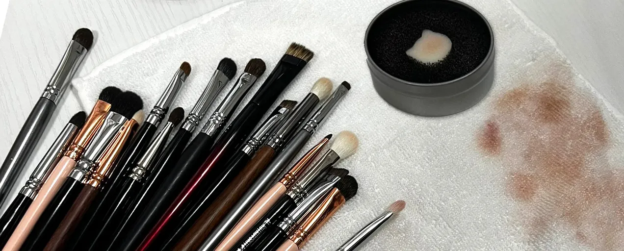 Makeup brushes laid out on a towel next to a dry cleaning sponge, used to quickly clean makeup brushes between clients and remove leftover color.
