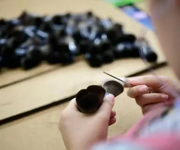 Worker hand‑shaping and trimming makeup brush heads with scissors to refine outline and layers.