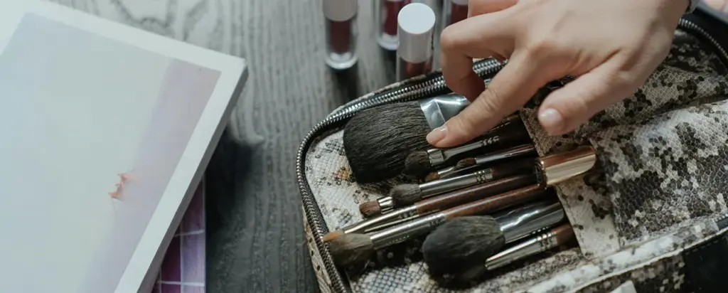 A hand choosing a makeup brush from a travel case filled with best quality makeup brushes on a dressing table.